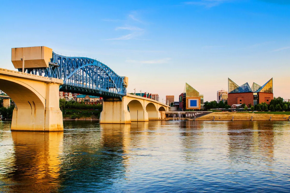 A scenic view of a blue bridge spanning a calm river, with modern architectural buildings in the background and the water reflecting the colors of sunset. The setting is tranquil and inviting, suggesting the nearby cityscape and opportunities for evening strolls or captivating sunset photography.