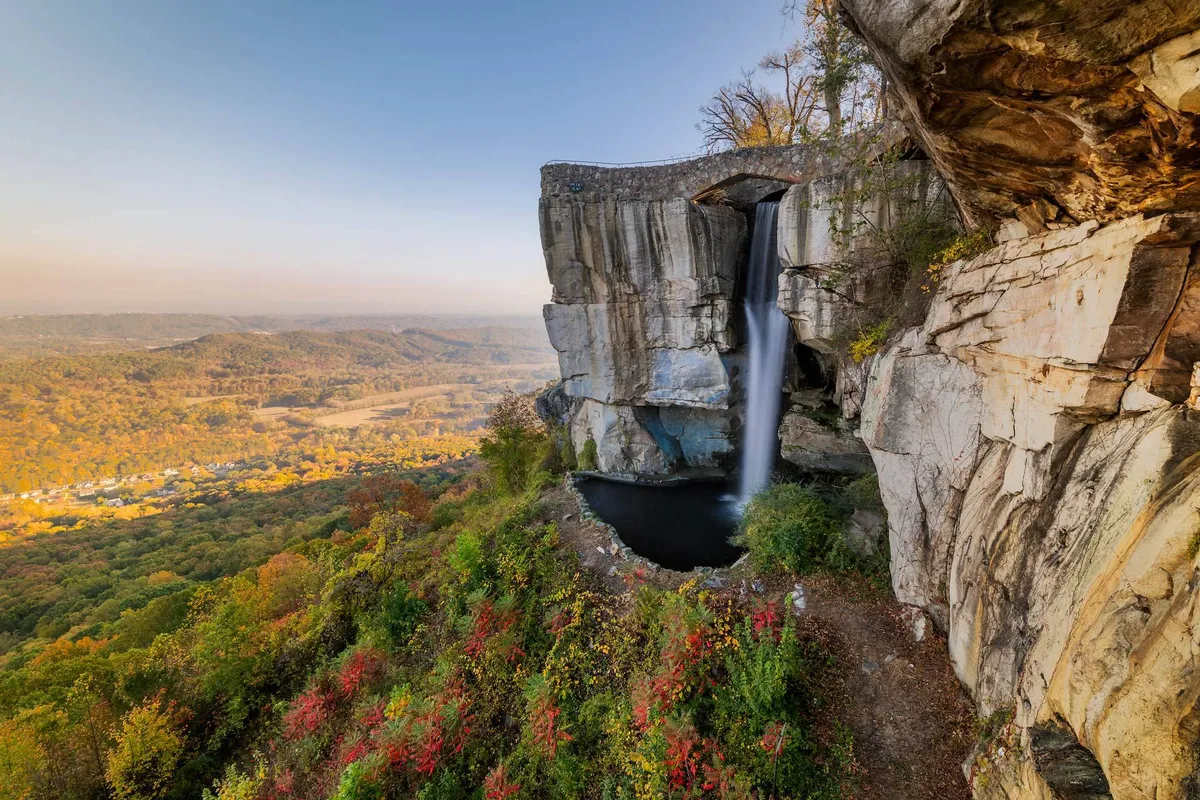 A dramatic exterior view of a cascading waterfall surrounded by rock formations, overlooking a lush valley with autumn foliage. The scene evokes a sense of natural wonder and outdoor adventure.
