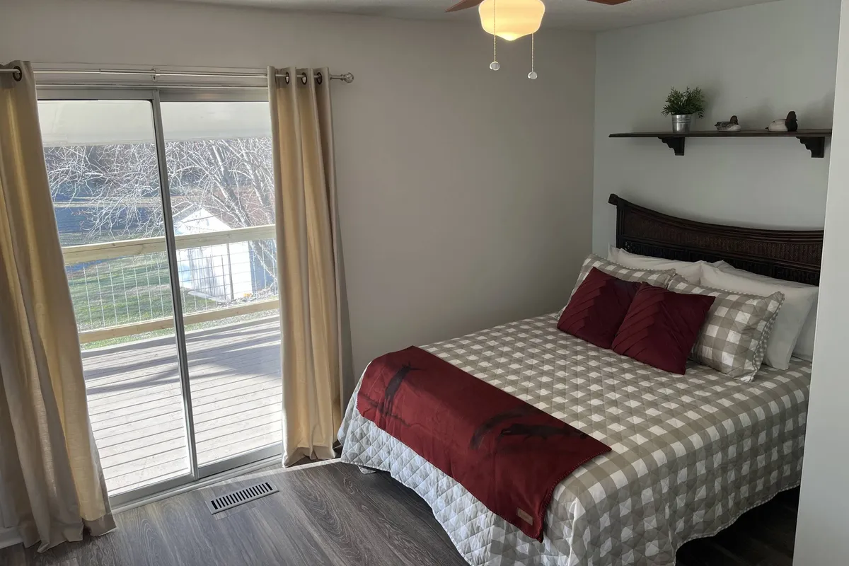 A cozy bedroom furnished with a bed featuring red and gray bedding, a shelf with decorative items, and a sliding glass door that leads out to a private deck. Natural light fills the room, creating a tranquil, inviting atmosphere.