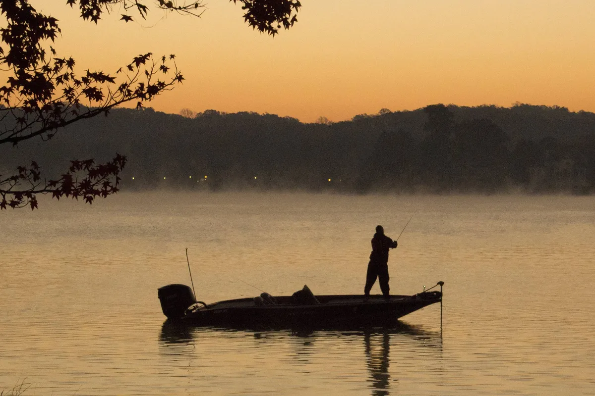 A fisherman stands on a boat in calm waters, framed by a stunning sunset sky. The scene captures the serenity and excitement of fishing at dusk, with the angler actively catching a fish, surrounded by peaceful nature.