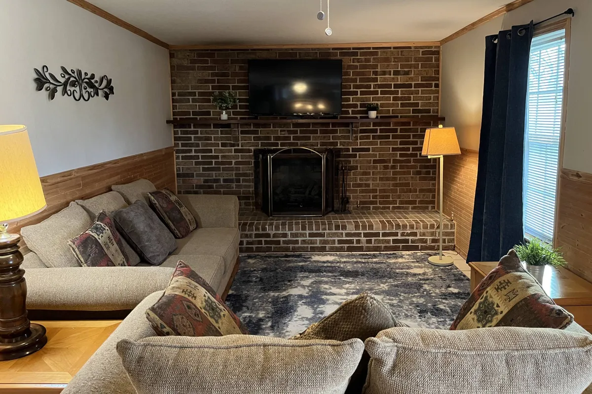 A spacious, inviting family den featuring a sofa, love seat, fireplace, television, rug, and lamps. The den offers a welcoming environment for family gatherings, game nights, or movie marathons.