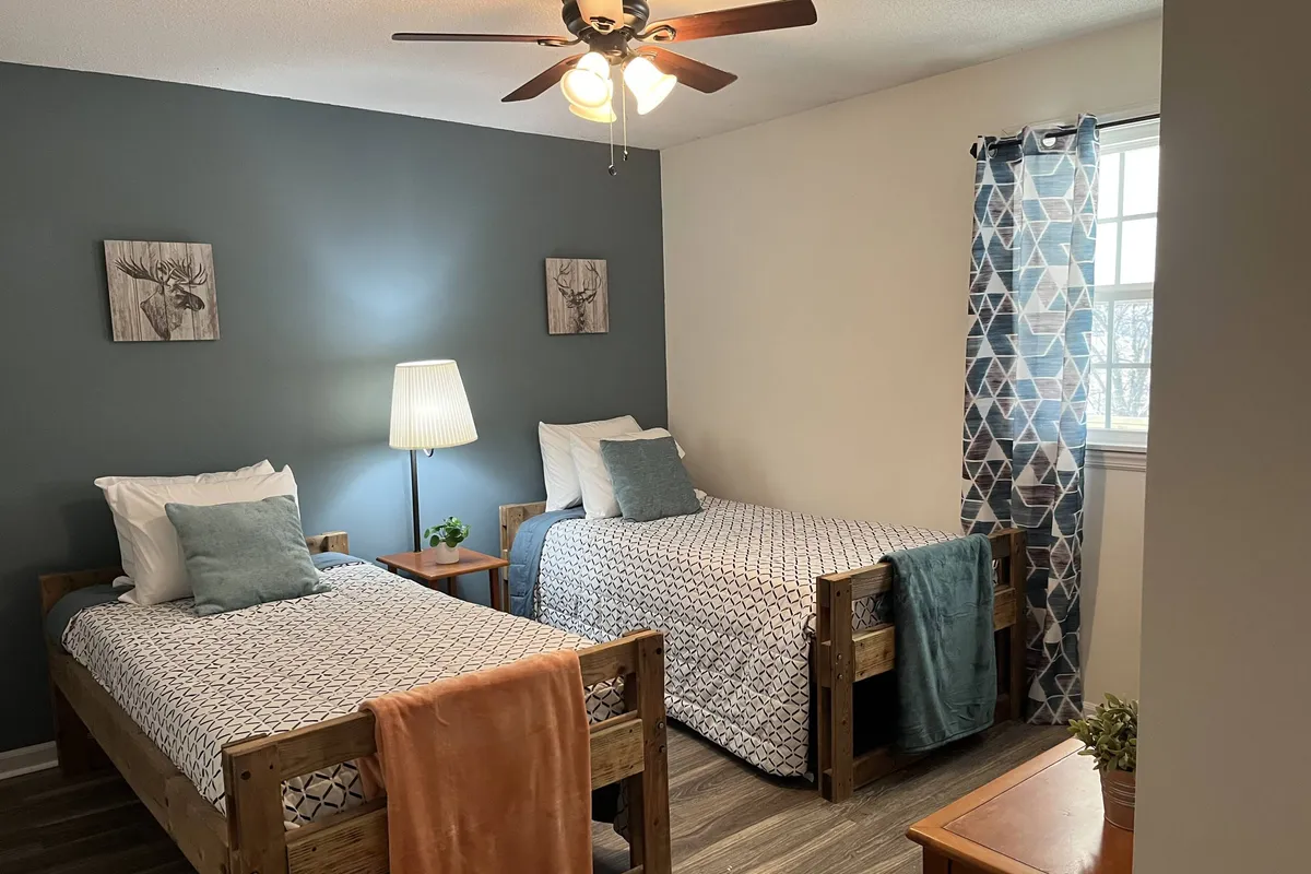 A comfortable bedroom with two twin beds, decorative wall art, a table lamp, and curtains. The setup encourages both quiet mornings and cozy conversations before bed, perfect for kids or friends sharing a room.