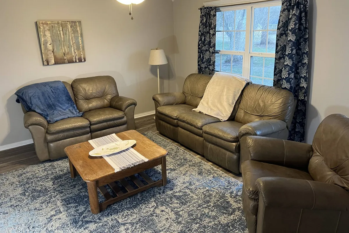 This image shows a cozy living room with leather sofas arranged around a wooden coffee table, decorative artwork on the walls, and a window with curtains letting in natural light. The space radiates comfort and is clearly designed for relaxation and socializing, with ample room for families or groups to gather after a day outdoors.