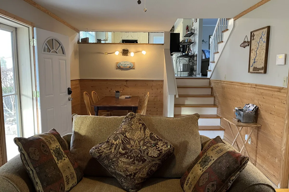 This interior view highlights a sofa adorned with decorative pillows, a dining table suitable for group meals or sorting fishing gear, a staircase indicating the tri-level layout, and tasteful wall decor. The open flow of the space connects various living areas, reinforcing a sense of spaciousness and utility for families and groups of anglers.