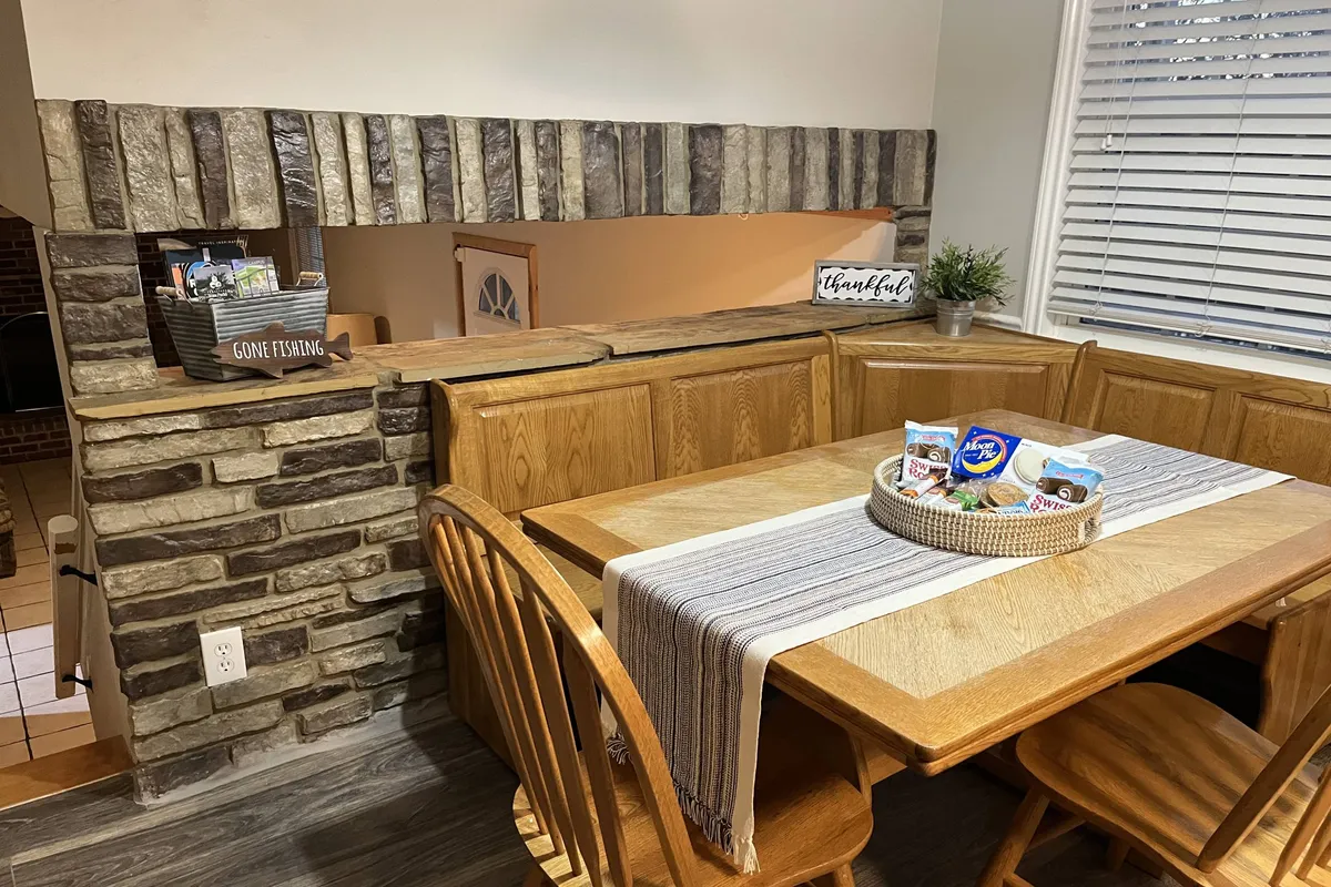 A rustic wooden dining table set beside a striking stone wall feature, with shelves displaying decorative items. The scene is intimate and inviting, perfect for family meals, celebration dinners, or game nights, all reinforcing the dining area's role as a communal hub.